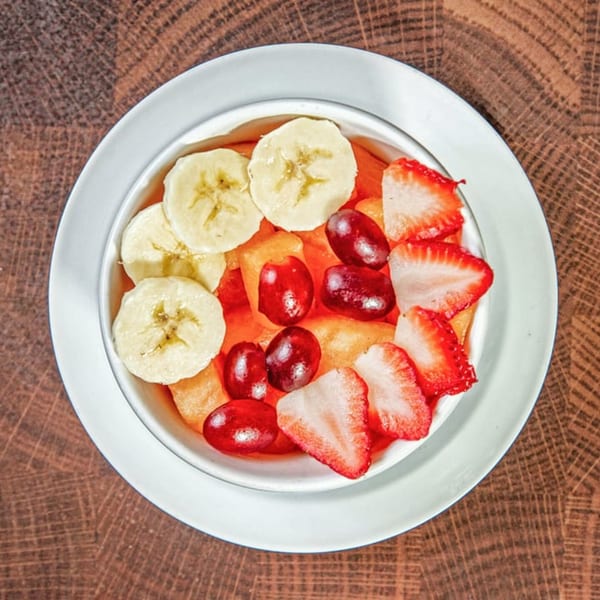 A white bowl of fresh fruit salad with bananas, strawberries, grapes, and melon on a wooden table.