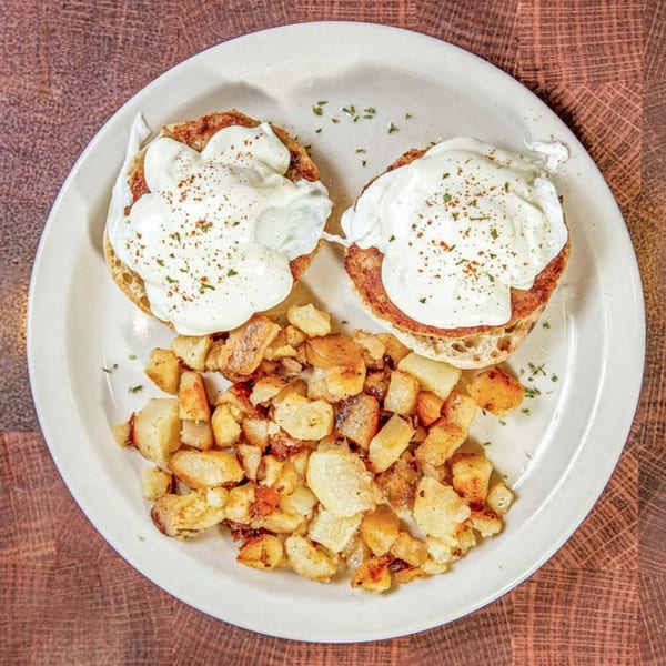 Two eggs benedict with hollandaise sauce and a side of crispy home fries on a white plate.